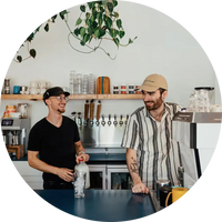 Two men standing in a kitchen, one holding a bottle, with a bar setup in the background.