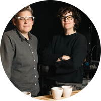 Two people standing behind a counter with coffee cups, wearing glasses.