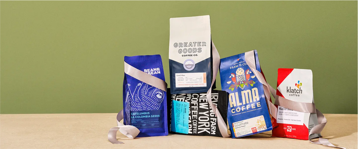 Five coffee bags from Trade roasters, wrapped in gray ribbon, stacked on a tan surface against an olive green wall