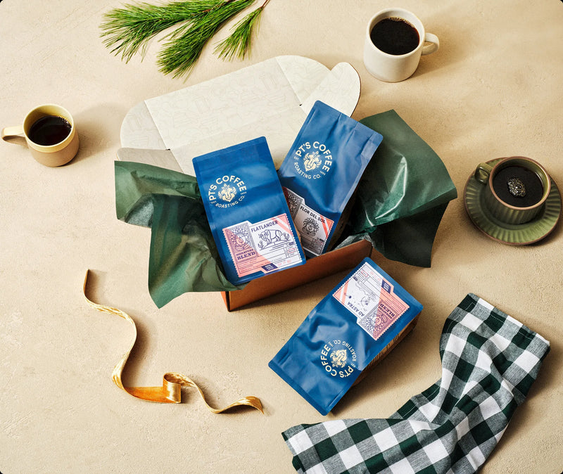 Open gift box with three bags of coffee, a pine sprig and a plaid napkin
