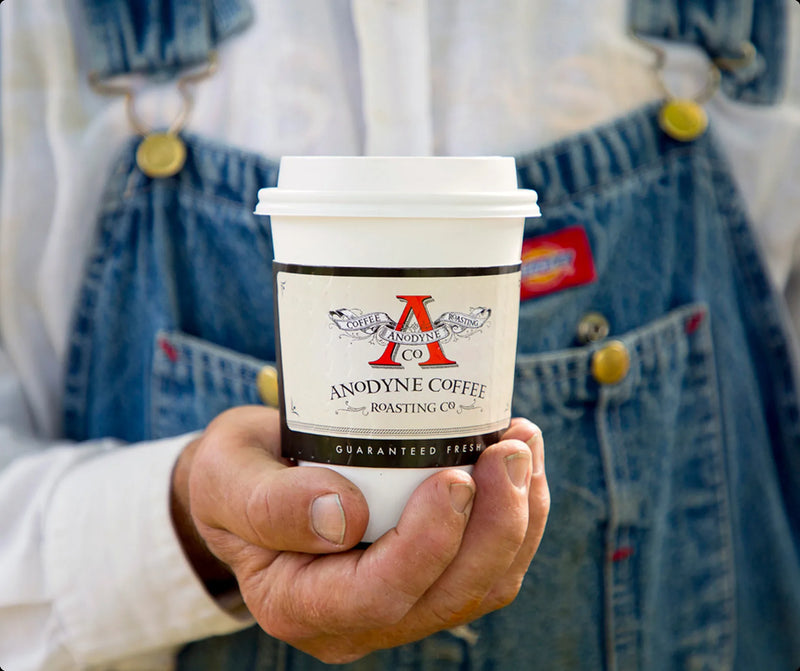 Hand holding a white Anodyne Coffee Roasting Co. cup in front of denim overalls