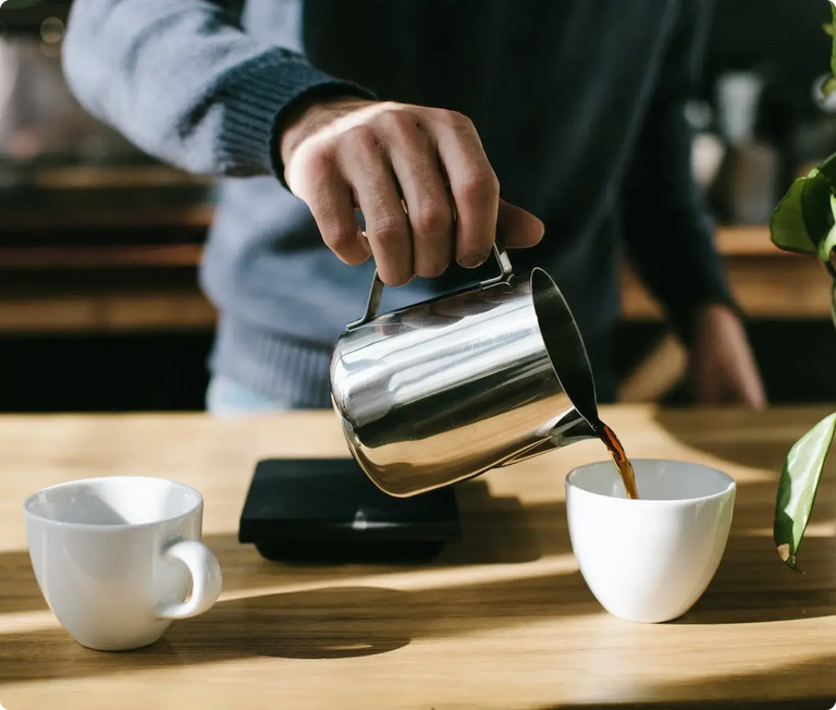 Person pouring coffee from a stainless steel pitcher into a white cup on a wooden table.